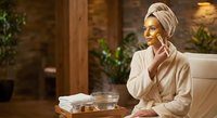 Young woman applying golden face mask in spa bathrobe and towel turban enjoying luxury skincare routine at home wellness retreat with natural lighting.
