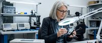 Senior female engineer with white hair and glasses examining robotic component in laboratory workshop with computers and equipment.