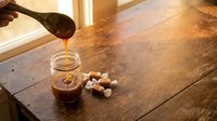 Golden honey being poured from wooden dipper into glass jar on rustic wooden table with scattered nuts creating warm natural kitchen scene.