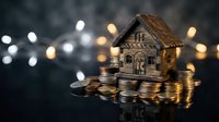 Miniature wooden house model sitting on stacks of coins with warm bokeh lights in background representing real estate investment and savings.