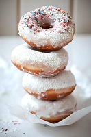 Stack of fresh glazed donuts with white frosting and colorful sprinkles on top, served on parchment paper. Close-up view shows soft texture and powdered sugar coating.