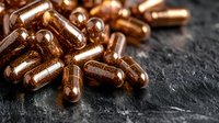 Shiny copper-colored supplement capsules scattered on dark stone surface, macro photography with selective focus and dramatic lighting for health products.