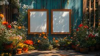 Empty wooden frames on rustic turquoise wall surrounded by vibrant garden flowers in terracotta pots, with dappled sunlight creating shadow patterns.