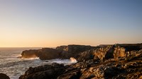 Dramatic rocky coastline with crashing waves at golden hour sunset, rugged cliffs and ocean spray creating scenic seascape for travel and nature content.