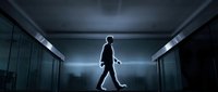 Silhouette of businessman walking through modern office corridor with dramatic lighting and glass walls creating professional corporate atmosphere.