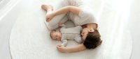 Young mother in beige clothing lying on woven rug holding newborn baby in gray outfit, bonding moment for family lifestyle and parenting content.