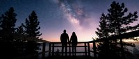 Silhouetted couple standing on wooden deck railing against starry night sky with glowing milky way galaxy and purple pink sunset horizon surrounded by evergreen trees.