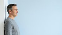 Middle-aged man with dark hair and stubble wearing gray casual shirt leaning against white wall in studio setting for portrait use.