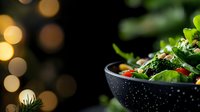 Fresh green salad with broccoli, cherry tomatoes and pine nuts served in black bowl on dark background with festive bokeh lights and copy space.