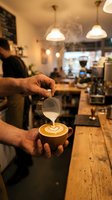 Barista pouring steamed milk into espresso cup creating latte art in modern cafe with warm pendant lighting and blurred background, suitable for coffee shop marketing and hospitality content.