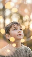 Curious Caucasian boy looking up with wonder, surrounded by golden bokeh lights in an autumn outdoor setting, warm sweater, dreamy mood.