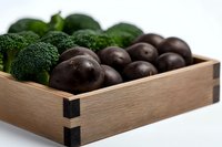 Fresh broccoli florets and dark purple potatoes arranged in rustic wooden box against white background, healthy organic produce for farm-to-table cooking.