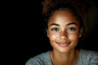 Young African American woman with natural curly hair and freckles smiling warmly at camera against dark background, wearing casual grey top. Genuine expression shows confidence.