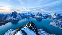 Majestic snow-capped mountain peaks surround crystal blue fjord waters in dramatic winter landscape, aerial view capturing pristine wilderness and serene Nordic scenery.