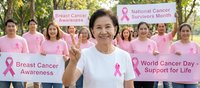 Senior Asian woman leading breast cancer awareness campaign with diverse group of female supporters holding pink ribbon signs in outdoor community setting.