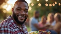 African American man smiling while holding grilled food at outdoor summer barbecue party with friends gathering in background during golden hour sunset.