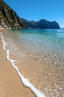 Pristine sandy beach with crystal clear turquoise water and dramatic coastal cliffs. Gentle waves wash onto golden sand creating natural patterns while rugged mountains rise from sea.