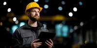 Confident male construction engineer in yellow hard hat using digital tablet at night construction site with bokeh lights in background.