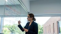 Professional businesswoman presenting mathematical formulas on whiteboard in modern office conference room for corporate training session.