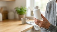 Senior woman holding white supplement bottle in kitchen with natural light and potted plant on wooden counter.