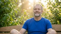 Middle-aged Caucasian man with gray beard wearing blue t-shirt sitting on wooden bench outdoors surrounded by green foliage and golden sunlight, suitable for lifestyle and wellness content.