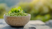 Fresh green peas in rustic stone bowl garnished with celery leaves on natural rock surface against blurred garden background. Organic healthy food concept.