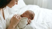 Young Caucasian mother in white clothing tenderly holding her newborn baby wrapped in knitted blanket on bed. Intimate bonding moment between parent and child.