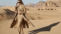 Woman in beige trench coat and sunglasses standing on sand dunes in desert landscape with camels in background. Adventure travel fashion.