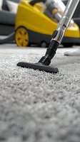 Close-up view of vacuum cleaner brush head on gray carpet with yellow vacuum cleaner in background, showing professional cleaning service in action.