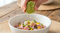 Hand squeezing fresh lime juice over colorful ceviche salad with diced fish, vegetables, and citrus in white bowl on wooden table.
