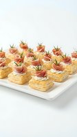 Elegant puff pastry appetizers topped with prosciutto and fresh rosemary on white serving platter for catering events and party entertaining.