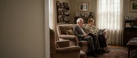 Elderly couple reading books together on comfortable armchair in cozy living room with warm lighting and family photos on shelves.