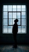 Silhouette of contemplative man standing by large window with grid pattern looking upward in moody indoor setting with natural light and wooden floor.
