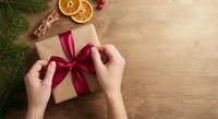 Hands wrapping Christmas gift with red ribbon on wooden table with festive decorations, dried orange slices and pine branches for holiday preparation.