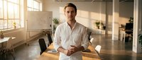 Male entrepreneur in white shirt standing at wooden desk in modern office space with natural sunlight, plants, and whiteboard, representing business leadership and professional workspace.
