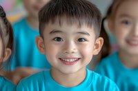 Young Asian boy wearing turquoise t-shirt smiling brightly at camera with genuine expression, close-up portrait against blurred children background in classroom setting.