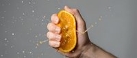 Hand squeezing fresh orange slice with juice splashing against neutral background, representing citrus freshness, healthy eating, and natural vitamin C source.