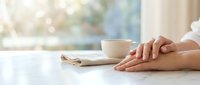 Woman hands resting on white table next to coffee cup in bright morning light with soft bokeh background creating peaceful atmosphere for relaxation and mindfulness.