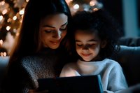 Young Asian mother and mixed race daughter smiling while looking at digital tablet screen in dark room illuminated by warm holiday lights, creating cozy atmosphere.
