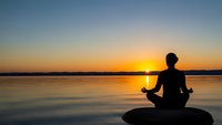 Person meditating in lotus position by calm lake at golden sunset with peaceful water reflections and serene sky for wellness and mindfulness concepts.