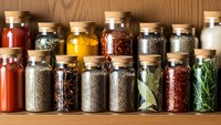 Collection of colorful spices and herbs in glass jars with cork lids arranged on wooden shelf for cooking and culinary seasoning purposes.