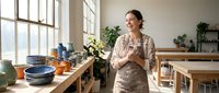 Smiling young Caucasian woman in apron stands in bright pottery studio surrounded by handmade ceramic bowls and vases, holding a mug.