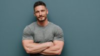 Confident muscular man with crossed arms smiling at camera wearing gray t-shirt against blue background for fitness and lifestyle marketing campaigns.