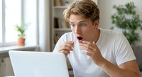 Young Caucasian man looking shocked while using laptop computer at home office desk, expressing surprise and amazement at screen content in modern interior.