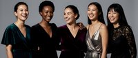 Five diverse women in elegant evening dresses laughing together against gray background, representing friendship, celebration and female empowerment.