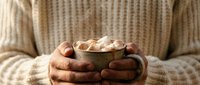 Cozy winter moment with hands holding warm mug of hot chocolate topped with marshmallows, wearing cream knit sweater for comfort and relaxation.