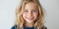 Young Caucasian girl with blonde wavy hair and bright blue eyes smiling at camera against white background, wearing blue sweater.