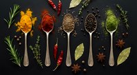 Colorful spices and herbs arranged in wooden spoons on dark background. Culinary ingredients including turmeric, paprika, cumin, peppercorns and herbs for cooking.