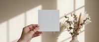 Hand holding a blank white square card mockup in soft natural light with dried flowers in background, perfect for branding.