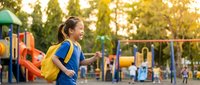 Happy young Caucasian girl with yellow backpack walking to school playground during golden hour sunset with colorful equipment in background for education concepts.